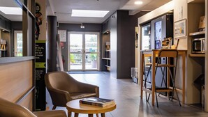A modern hotel lobby with leather seating, a small round table with magazines, a bar area with a computer and a refrigerator, and a large window offering a view of the outdoors. - B&B HOTEL Valence Sud (Valence)
