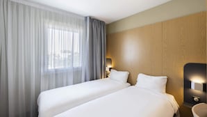A hotel room with two beds, a large window with sheer curtains, a wooden headboard, and a bedside table with a lamp. - B&B HOTEL Perpignan Sud Marché International (Perpignan)