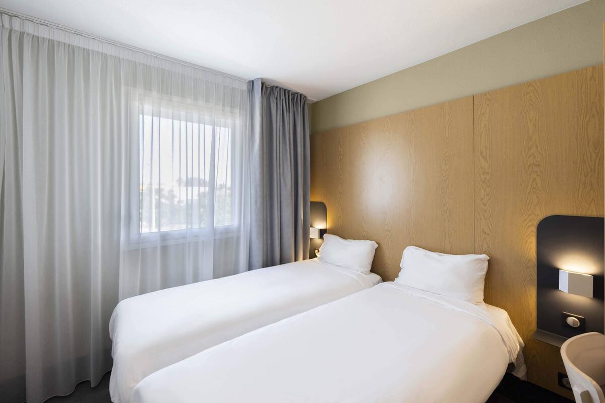 Habitación de hotel con dos camas, un ventanal con cortinas translúcidas, cabecera de madera y mesita de noche con lámpara.
