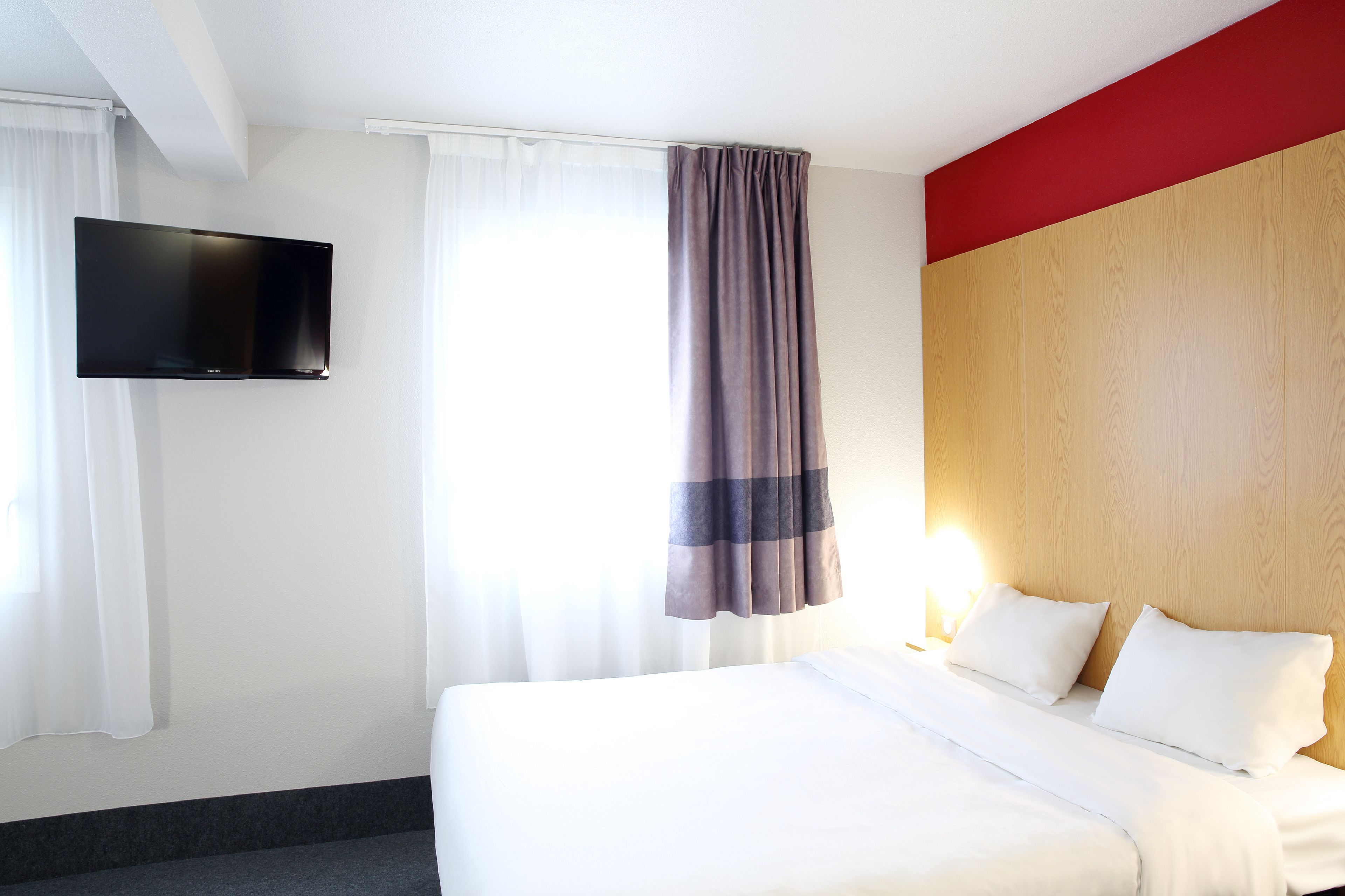 A hotel room with a bed, a flat-screen TV mounted on the wall, and a window with curtains.