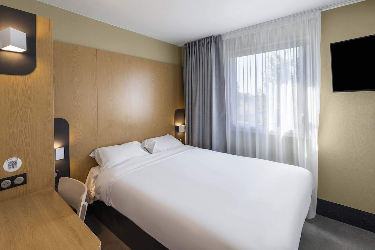 Habitación de hotel con una cama grande, un escritorio, una silla, un televisor de pantalla plana y una ventana con cortinas.