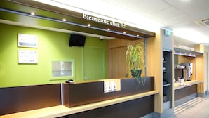 A reception desk with a 'Bienvenue chez' sign, a framed certificate, and a potted plant. - B&B HOTEL Perpignan Sud Marché International (Perpignan)