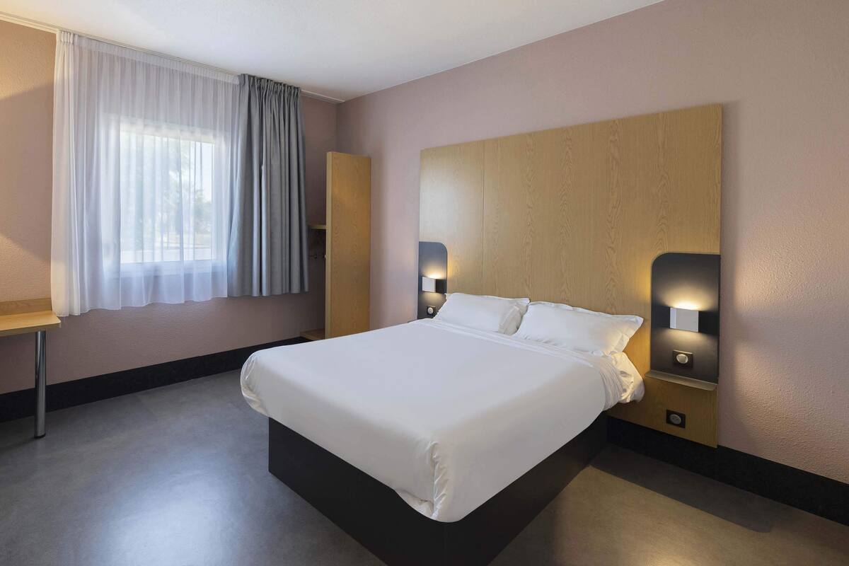 Una habitación de hotel con una cama grande, una mesita de noche, una lámpara y una ventana con cortinas.
