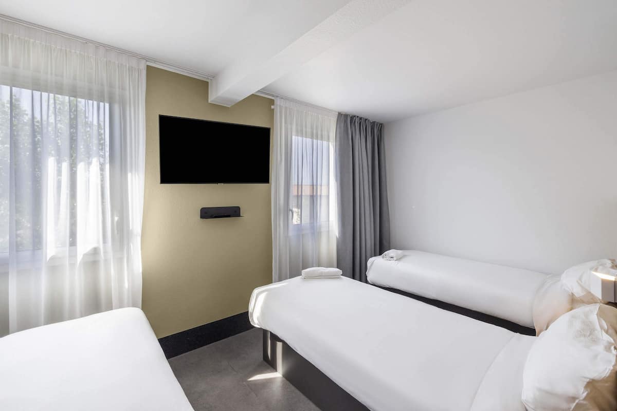 Habitación de hotel con dos camas, un televisor de pantalla plana montado en la pared y cortinas translúcidas en las ventanas.