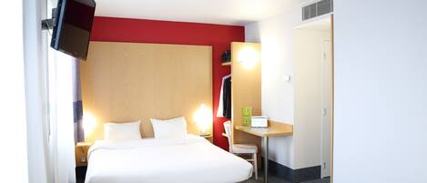 A hotel room with a bed, a desk, a chair, a TV, and a door.