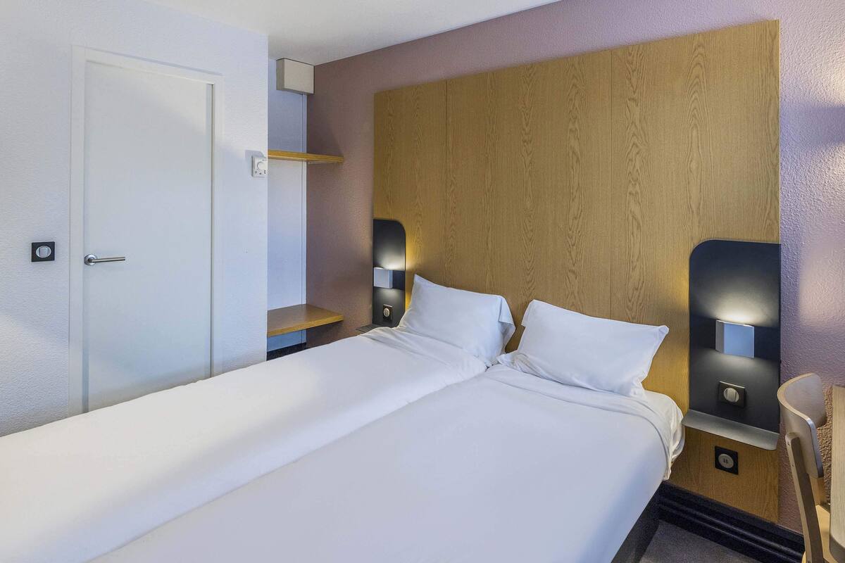 Habitación de hotel con una cama grande, dos mesitas de noche con lámparas, un cabecero de madera, una puerta blanca y un pequeño estante de madera.