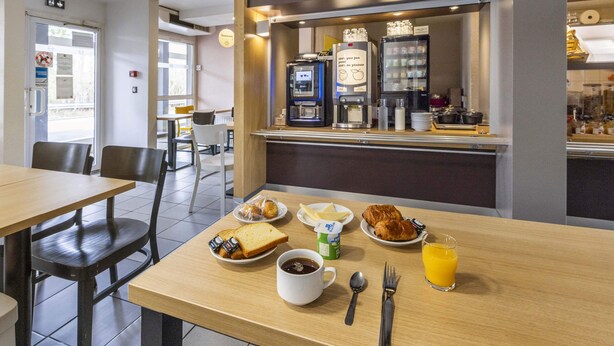 Un desayuno con una mesa preparada para dos personas, que incluye croissants, tostadas, una taza de café, un vaso de jugo de naranja y un envase de yogur.