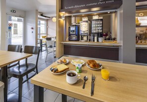 Une table mise pour le petit-déjeuner, avec une place pour deux personnes, comprenant des croissants, des toasts, une tasse de café, un verre de jus d’orange et un pot de yaourt.