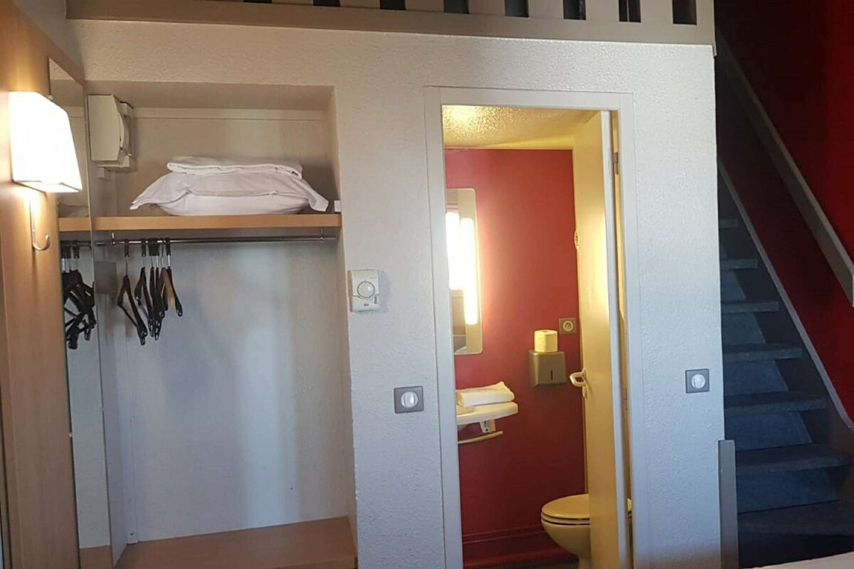 A hotel room with a bed, a closet with hanging clothes, a bathroom with a toilet, and a staircase.