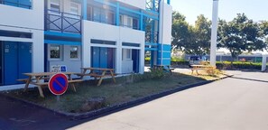 Edifício com fachada azul e branca, mesas de piquenique, placa de proibição de estacionamento e sinalização de acessibilidade para cadeiras de rodas.