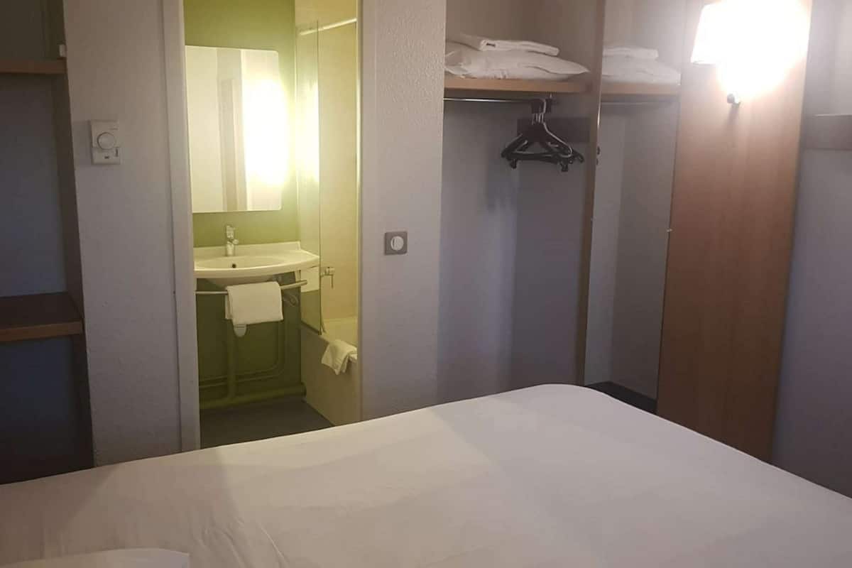 Habitación de hotel con cama, baño con lavamanos y espejo, y armario con ropa colgada.