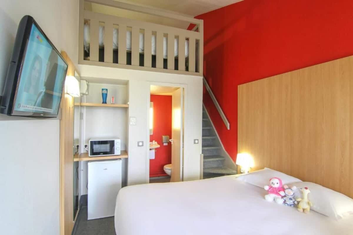 Habitación de hotel con una cama, un escritorio con microondas, televisor, una escalera y una pared roja.