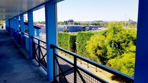A covered walkway with blue pillars and a metal railing, overlooking a parking area and greenery.