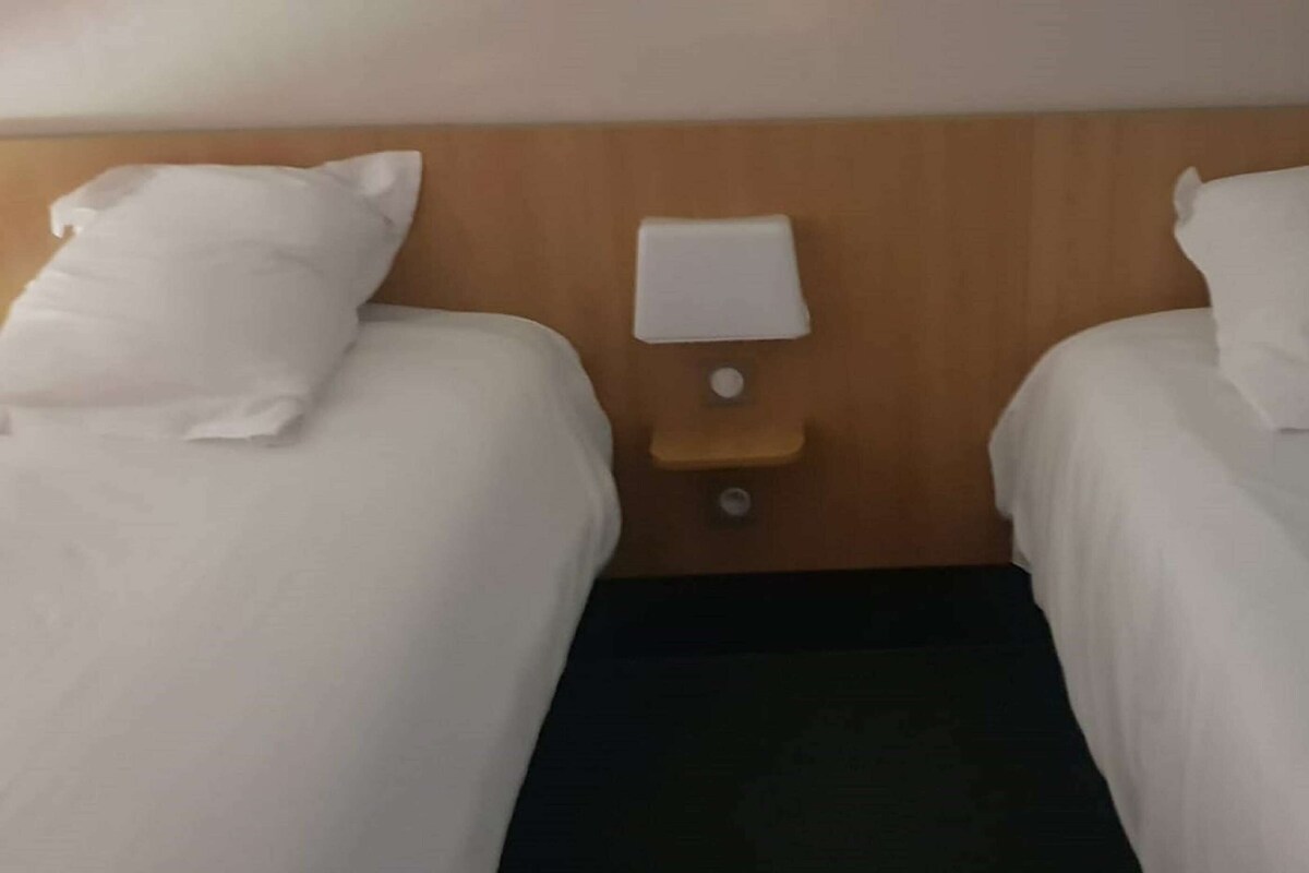A hotel room with two single beds, each with white linens, a wooden headboard, and a small bedside table with a lamp.
