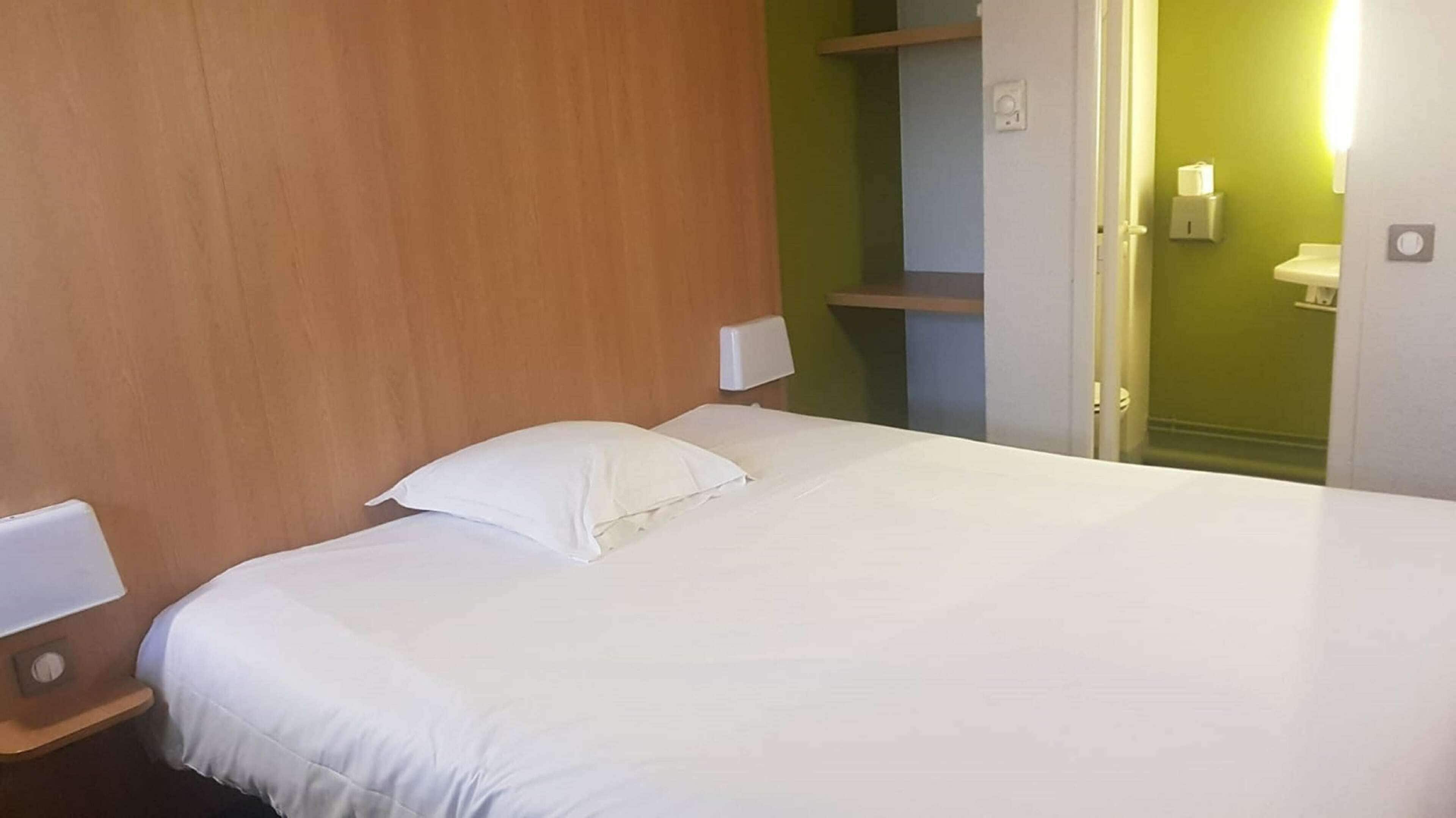 A hotel room with a wooden headboard, a bed with white linens, a bedside table with a lamp, and a green wall with a shelf.
