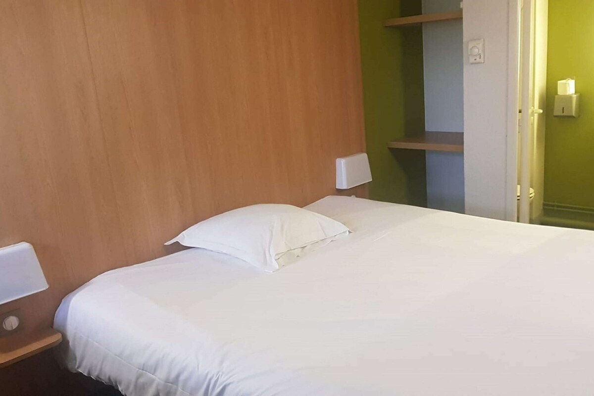 A hotel room with a wooden headboard, a bed with white linens, a bedside table with a lamp, and a green wall with a shelf.