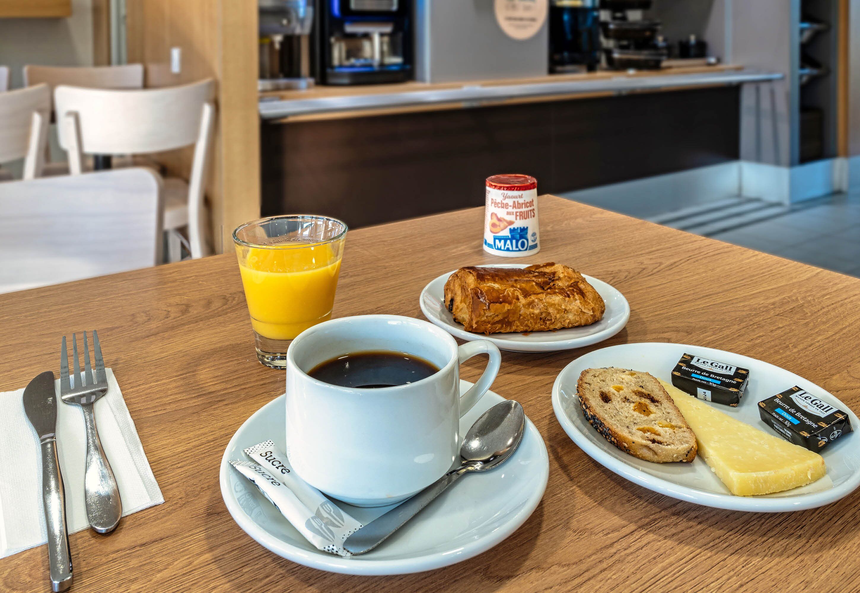 A breakfast setting with a cup of coffee, a glass of orange juice, a croissant, a slice of bread, cheese, and jam on a wooden table.