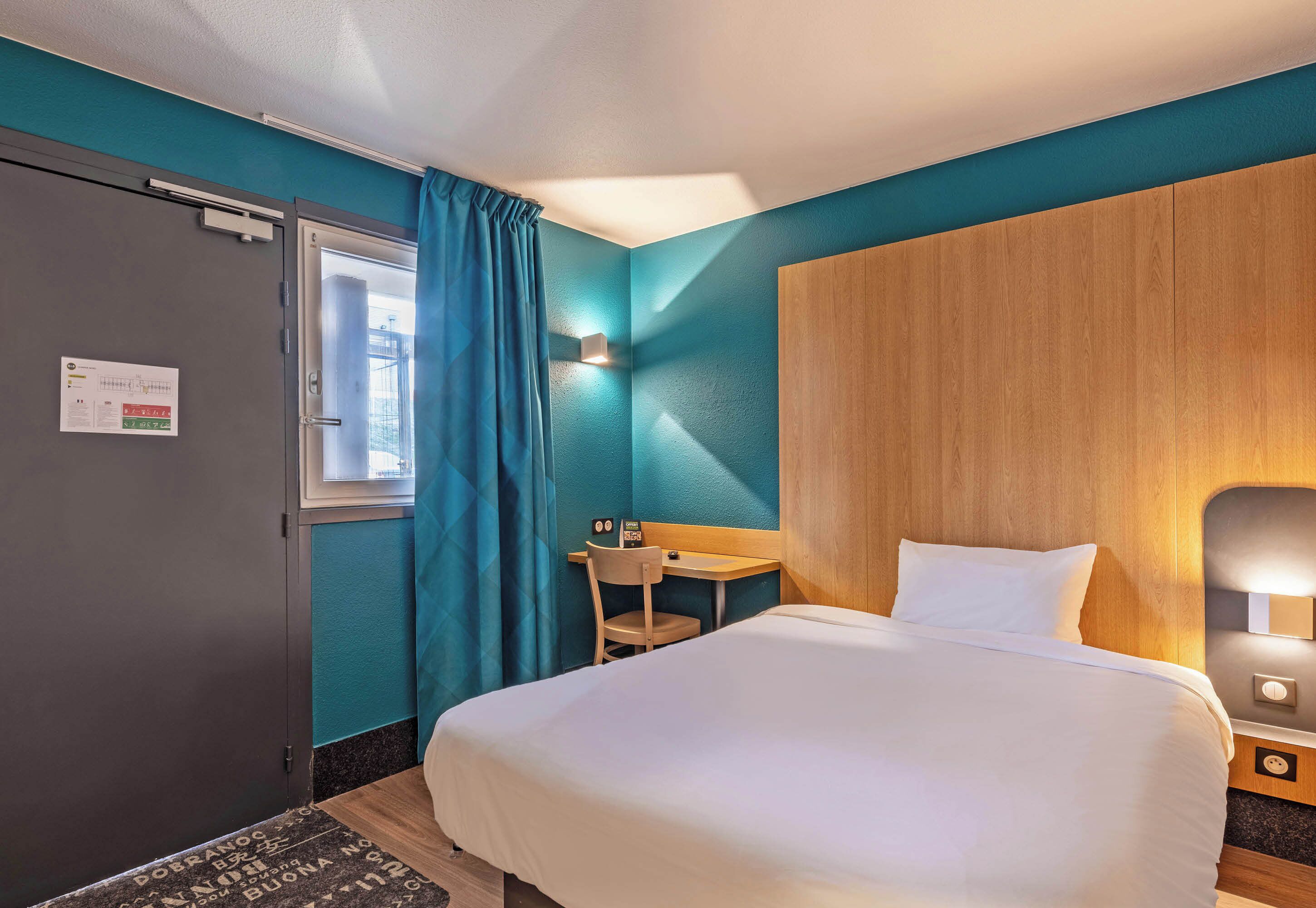 Quarto de hotel com cama, escrivaninha, cadeira e janela com cortinas.