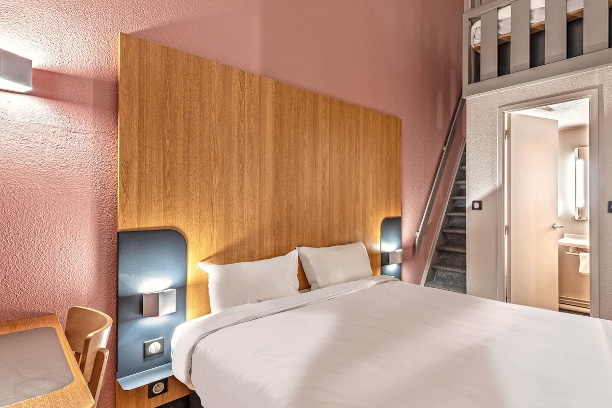 Een hotelkamer met een houten hoofdeinde, een bed met witte lakens, een nachtkastje met een lamp, en een trap naar een hoger gelegen niveau.