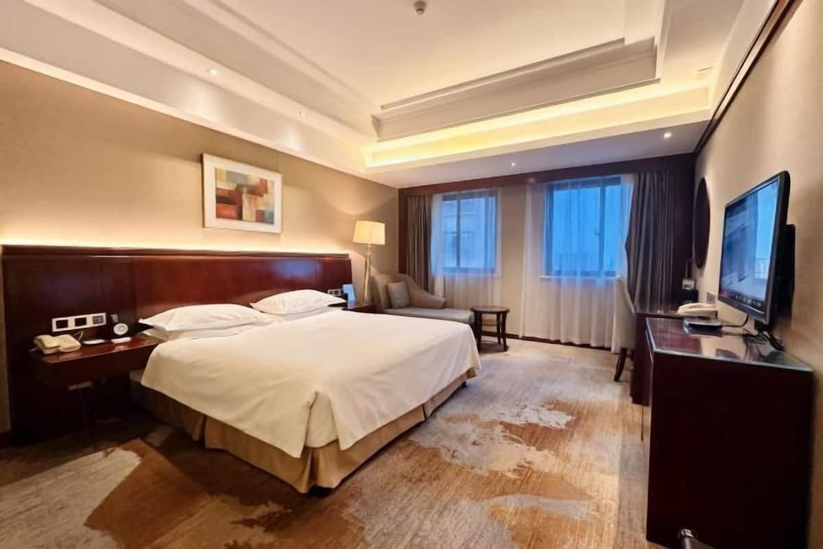 Une chambre d’hôtel avec un grand lit, un bureau avec une lampe, une télévision et une fenêtre avec des rideaux.
