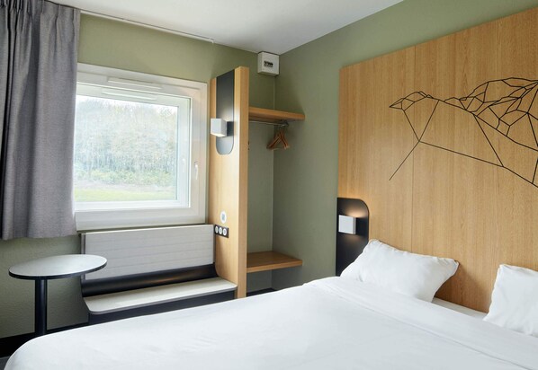 Une chambre d’hôtel avec un lit, une petite table, un porte-manteau et une fenêtre avec des rideaux.