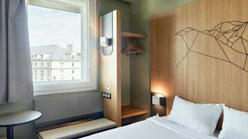 A hotel room with a bed, a bench, a small table, a coat rack, and a window with a view of buildings.