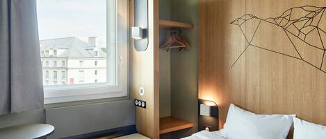 A hotel room with a bed, a bench, a small table, a coat rack, and a window with a view of buildings.
