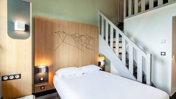 Une chambre d’hôtel moderne avec une tête de lit en bois, un lit recouvert de draps blancs, deux lampes de chevet, une table de chevet et un escalier avec des rampes blanches.