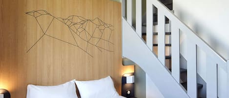 A hotel room with a wooden headboard, a bed with white bedding, two bedside lamps, and a staircase.