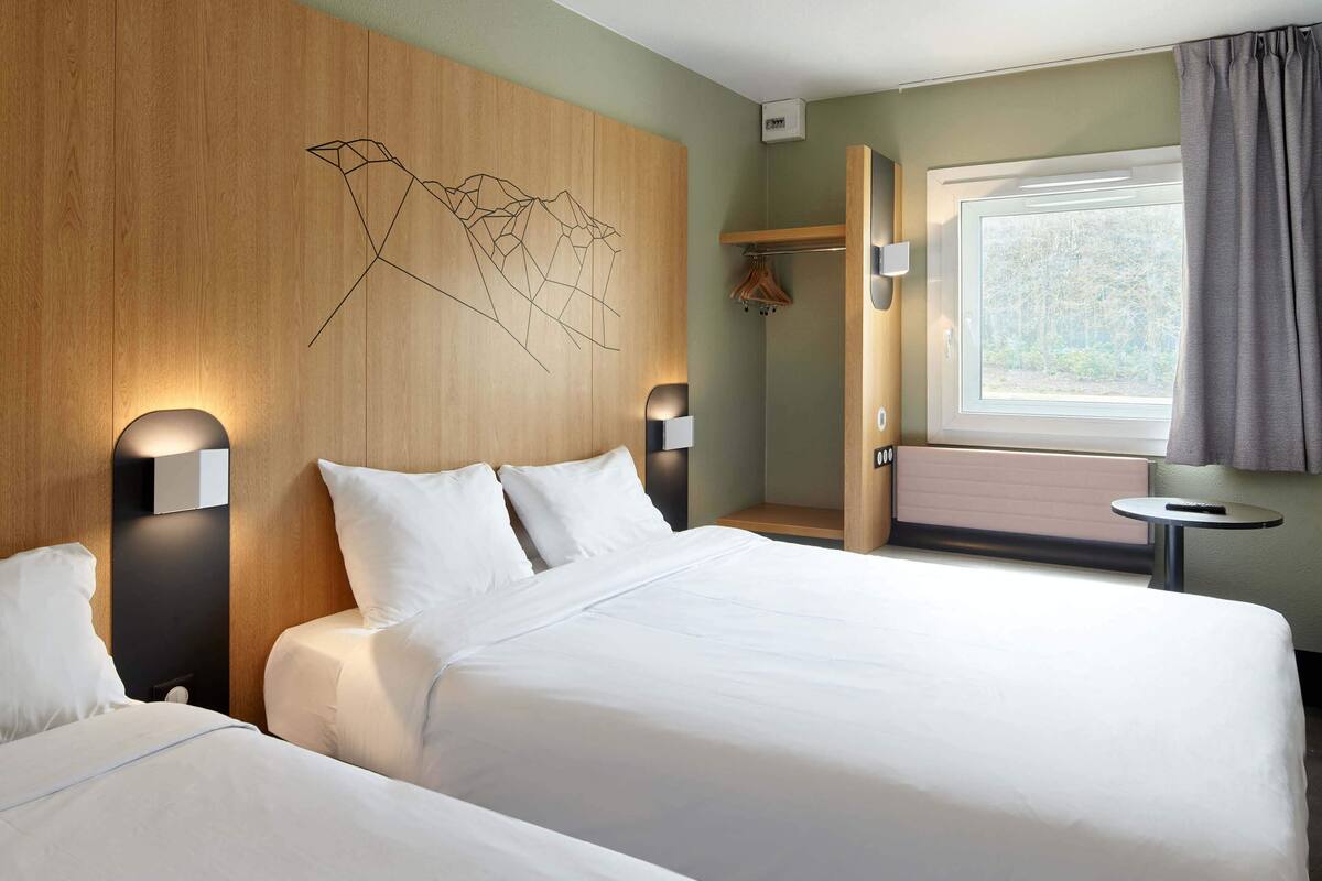 Ein Hotelzimmer mit zwei Betten, einem Holzkopfteil mit geometrischem Muster, einem Fenster mit Vorhängen und einem kleinen Tisch mit einer Fernbedienung.