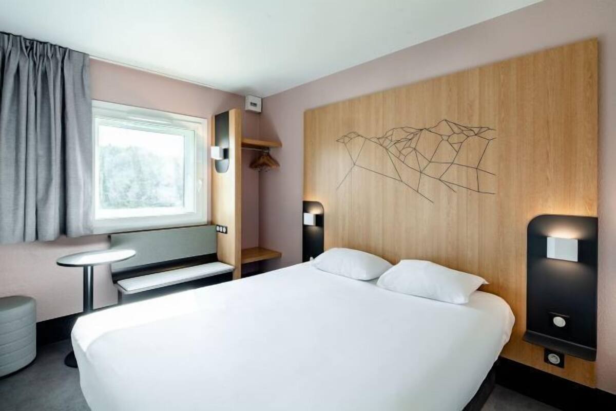 Une chambre d’hôtel moderne dotée d’un grand lit, de tables de chevet, d’un petit bureau, d’une fenêtre avec des rideaux et d’un mur décoratif en bois orné d’un motif géométrique.