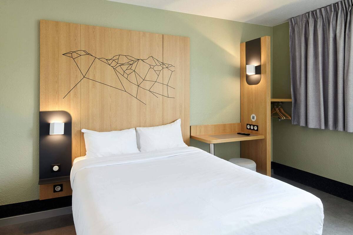 Ein Hotelzimmer mit einem großen Bett, einem hölzernen Kopfende mit geometrischem Muster, einem Schreibtisch mit Stuhl, einem Spiegel und einem Kleiderschrank.
