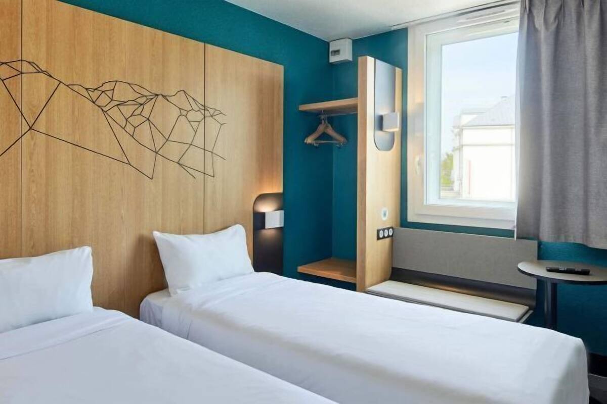Une chambre d’hôtel avec deux lits, une tête de lit en bois, une décoration murale aux motifs géométriques, une fenêtre avec des rideaux et une petite table.