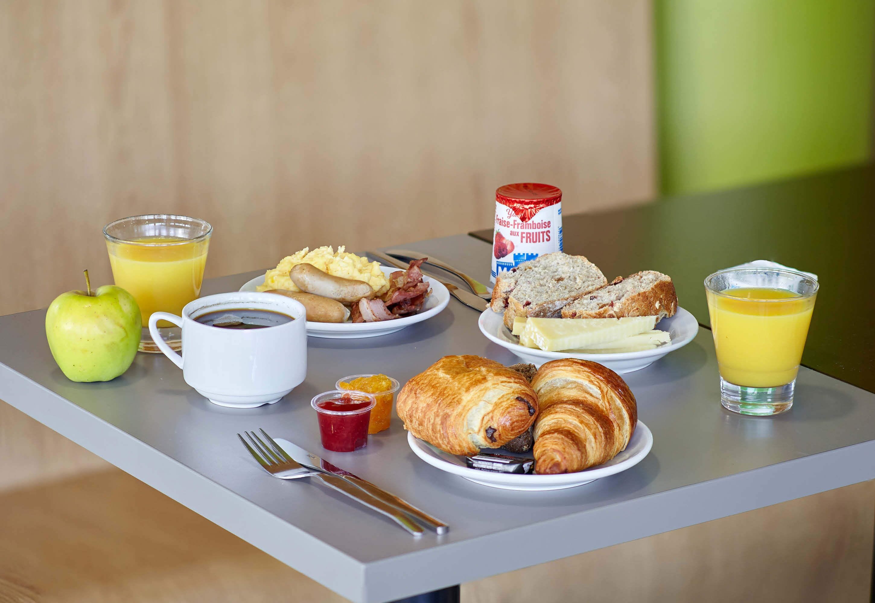 Une table de petit-déjeuner avec des croissants, des œufs, du bacon, un pot de confiture de fruits, un verre de jus d’orange, une tasse de café, une tranche de pain, une banane et une pomme verte.