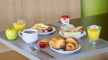 A breakfast table with croissants, eggs, bacon, a can of fruit spread, a glass of orange juice, a cup of coffee, a slice of bread, a banana, and a green apple.