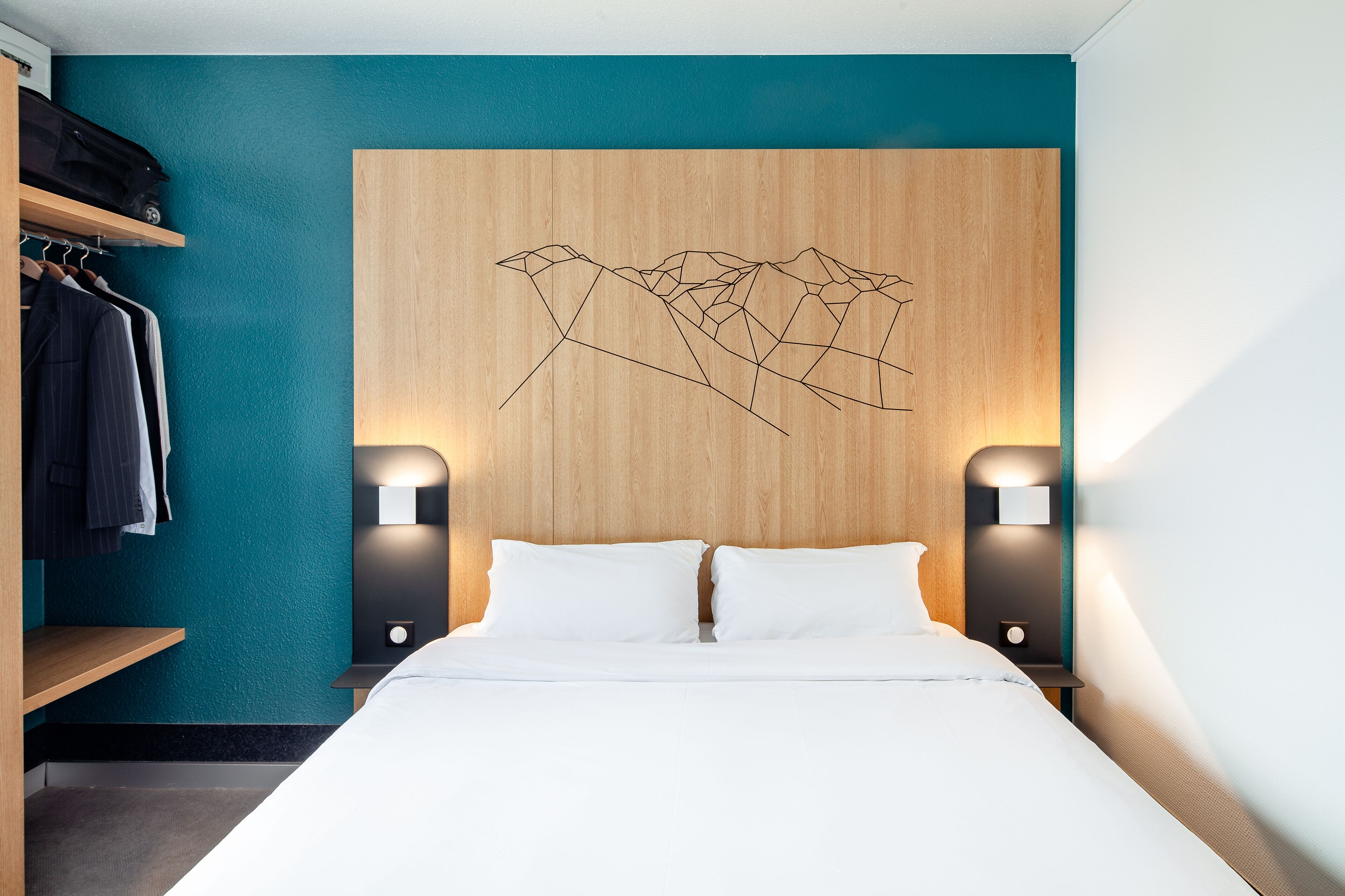 A modern hotel room with a wooden headboard, a teal accent wall, and a geometric wall art piece.