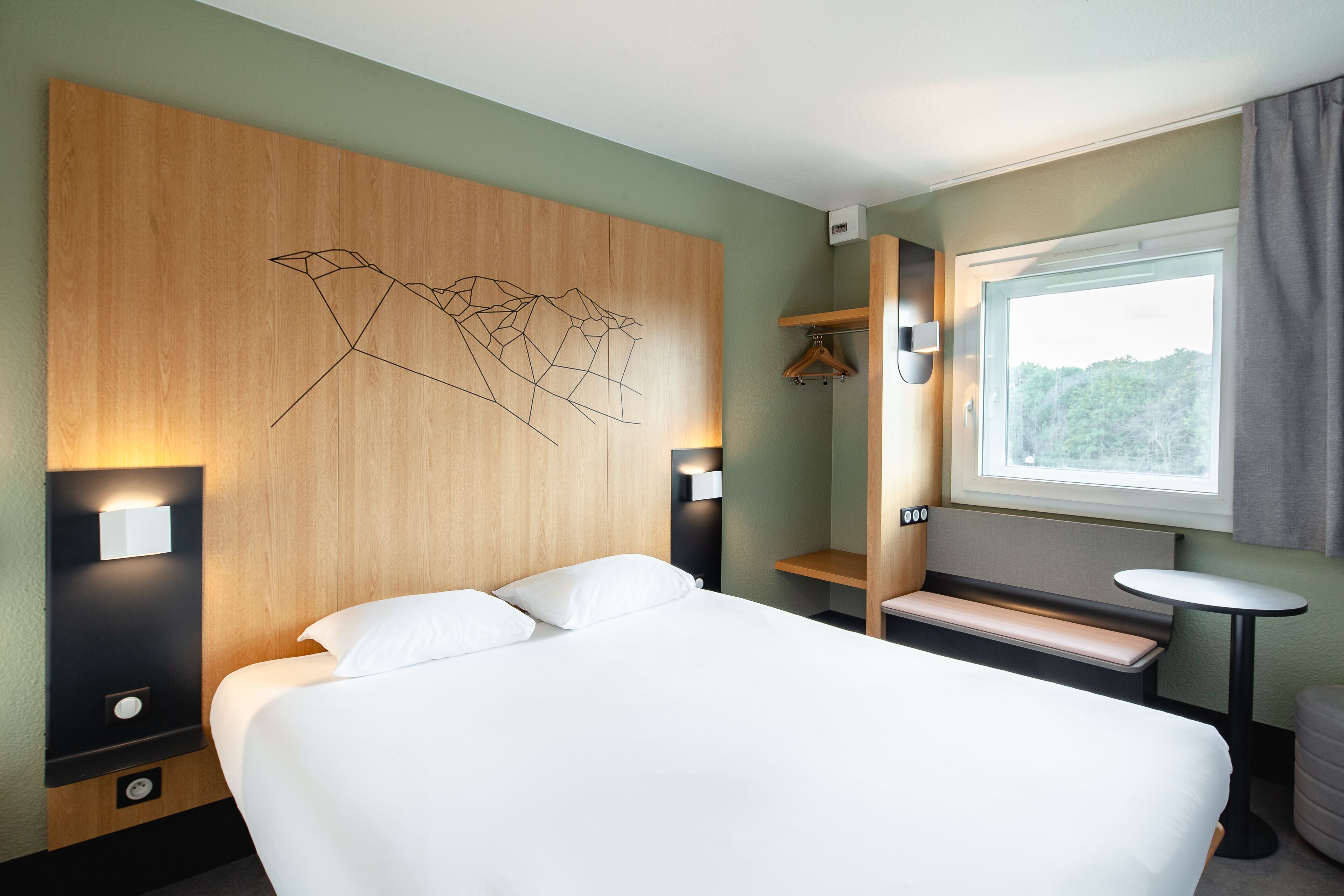Quarto de hotel moderno com uma cama grande, cabeceira de madeira com design geométrico, mesas de cabeceira, um banquinho, uma pequena mesa redonda e uma janela com vista para vegetação.