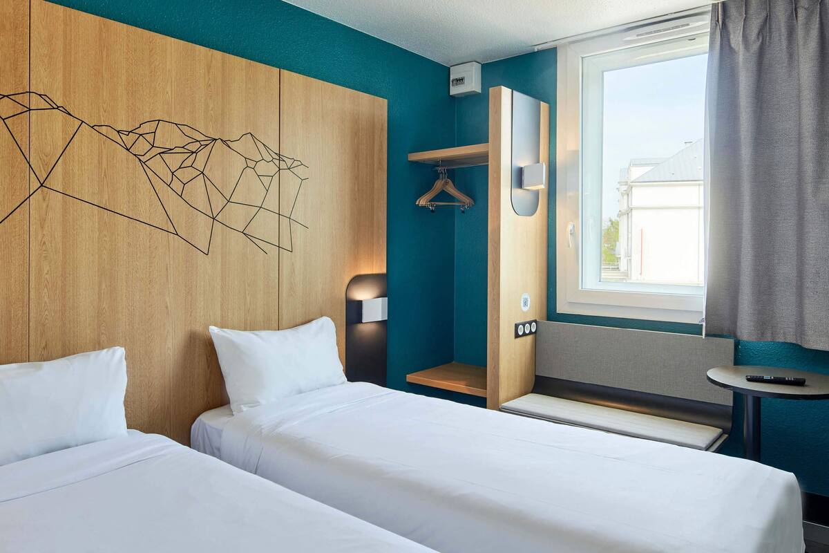 Ein Hotelzimmer mit zwei Betten, einem Holzkopfende, einem Wandbild, einem Nachttisch, einem Hängekleiderständer und einem Fenster mit Blick auf Gebäude.