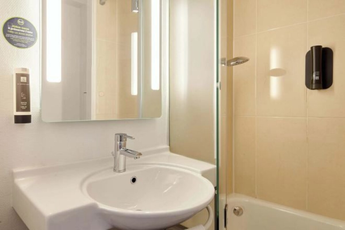 Une salle de bain avec un lavabo blanc, un miroir, une cabine de douche en verre et un distributeur de savon fixé au mur.