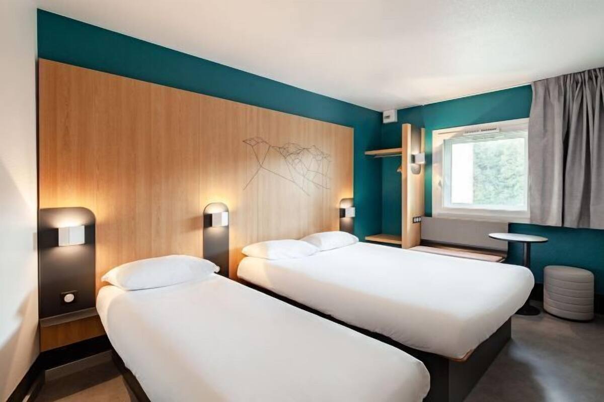 Une chambre d’hôtel avec deux lits, une tête de lit en bois, une fenêtre avec des rideaux et une petite table.