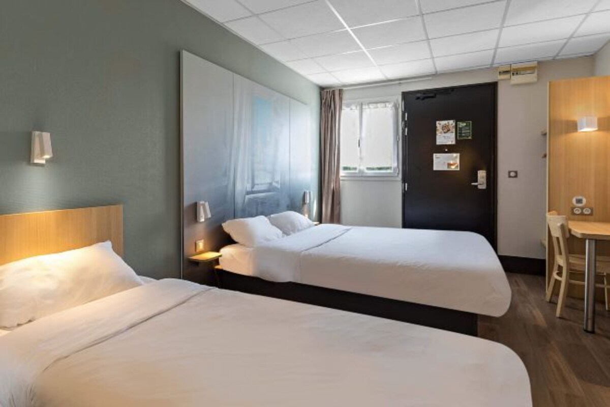 A hotel room with two beds, a large mirror, a window with curtains, a door with a sign, and a small table with a chair.