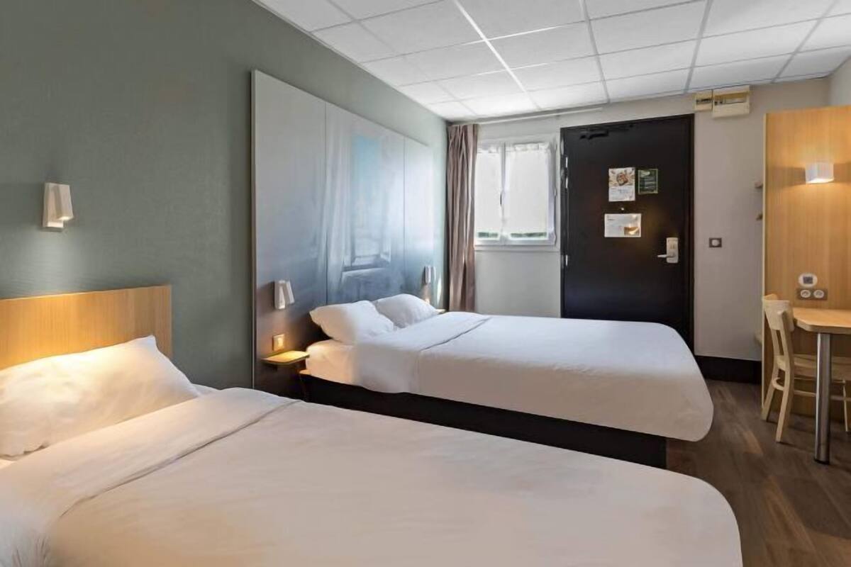 A hotel room with two beds, a large mirror, a window with curtains, a door with a sign, and a small table with a chair.