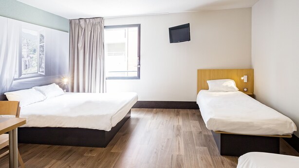 Une chambre d’hôtel avec deux lits, un bureau, une fenêtre avec des rideaux et un téléviseur à écran plat fixé au mur.