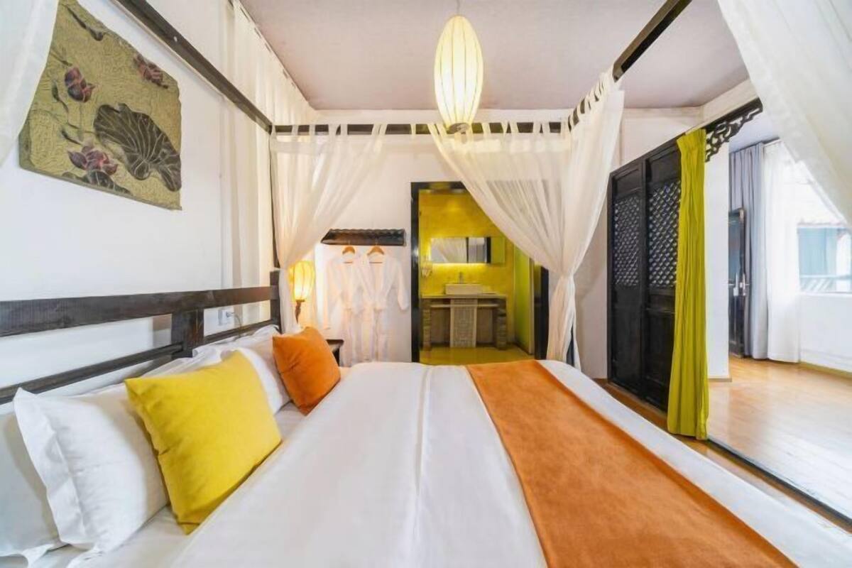 A bedroom with a bed, white curtains, a hanging light fixture, and a view of a bathroom.