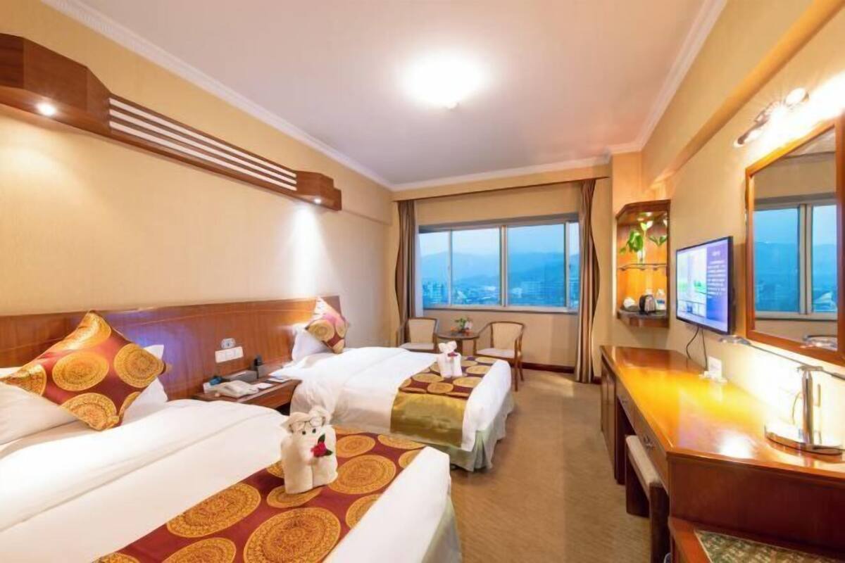 A hotel room with two beds, a desk, a TV, and a view of the city.