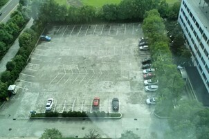 Vista aérea de un estacionamiento con múltiples filas de coches aparcados, rodeado de vegetación y un edificio en un lateral.