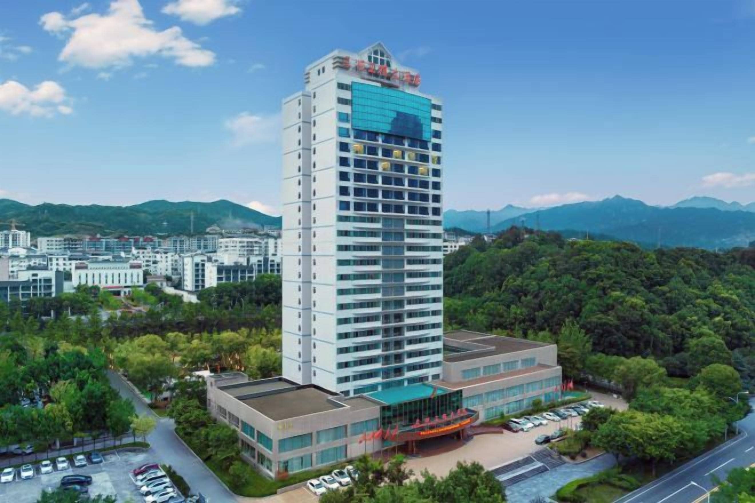 A high-rise hotel with multiple floors, a parking area, and a view of surrounding greenery and mountains.