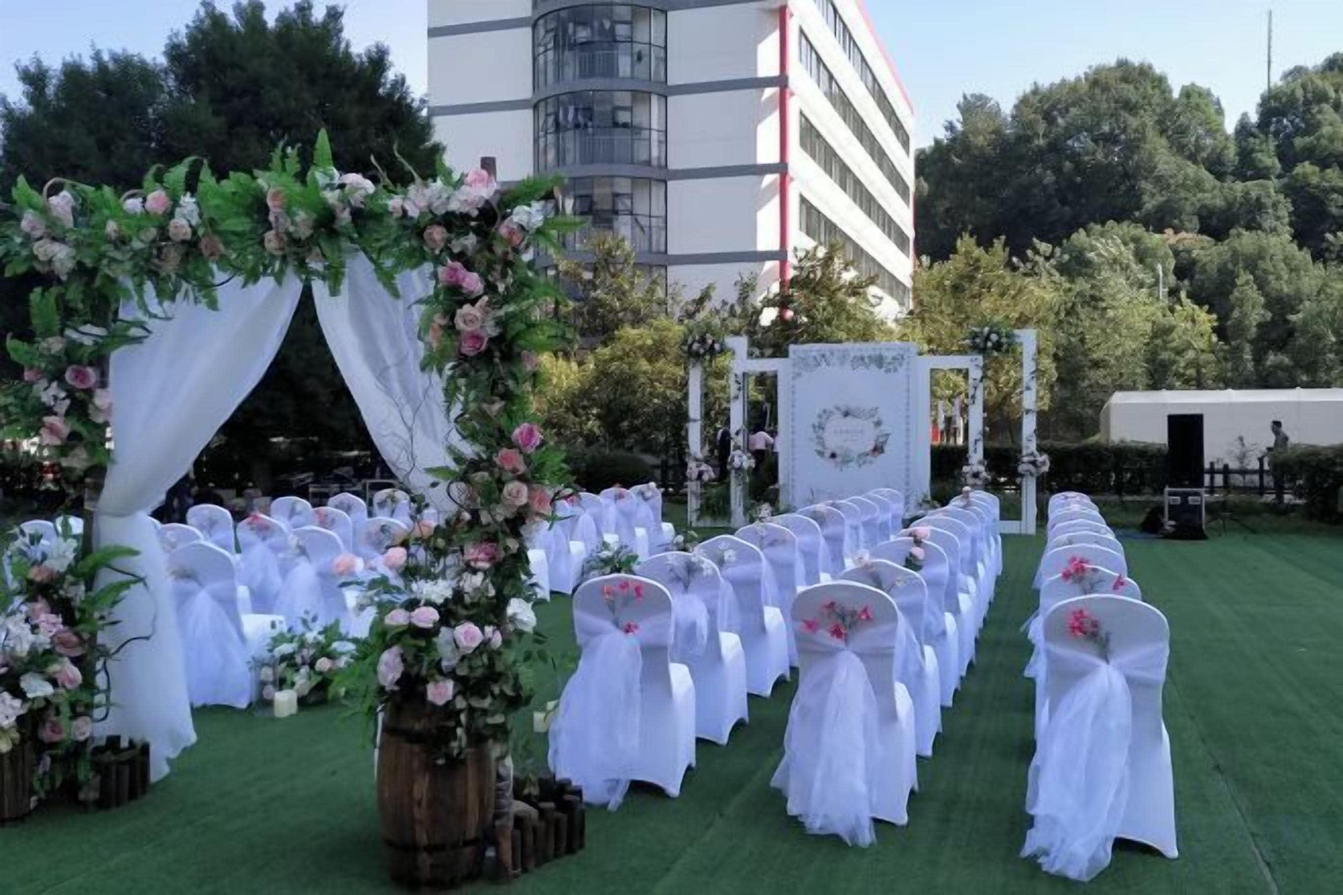 Salón de bodas al aire libre con filas de sillas blancas, un arco floral y un fondo con diseño circular.