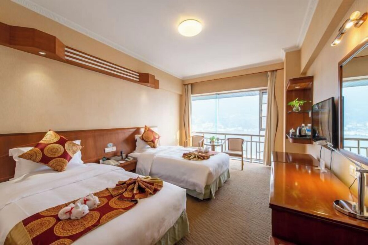A hotel room with two beds, a TV, a desk, and a balcony with a view.