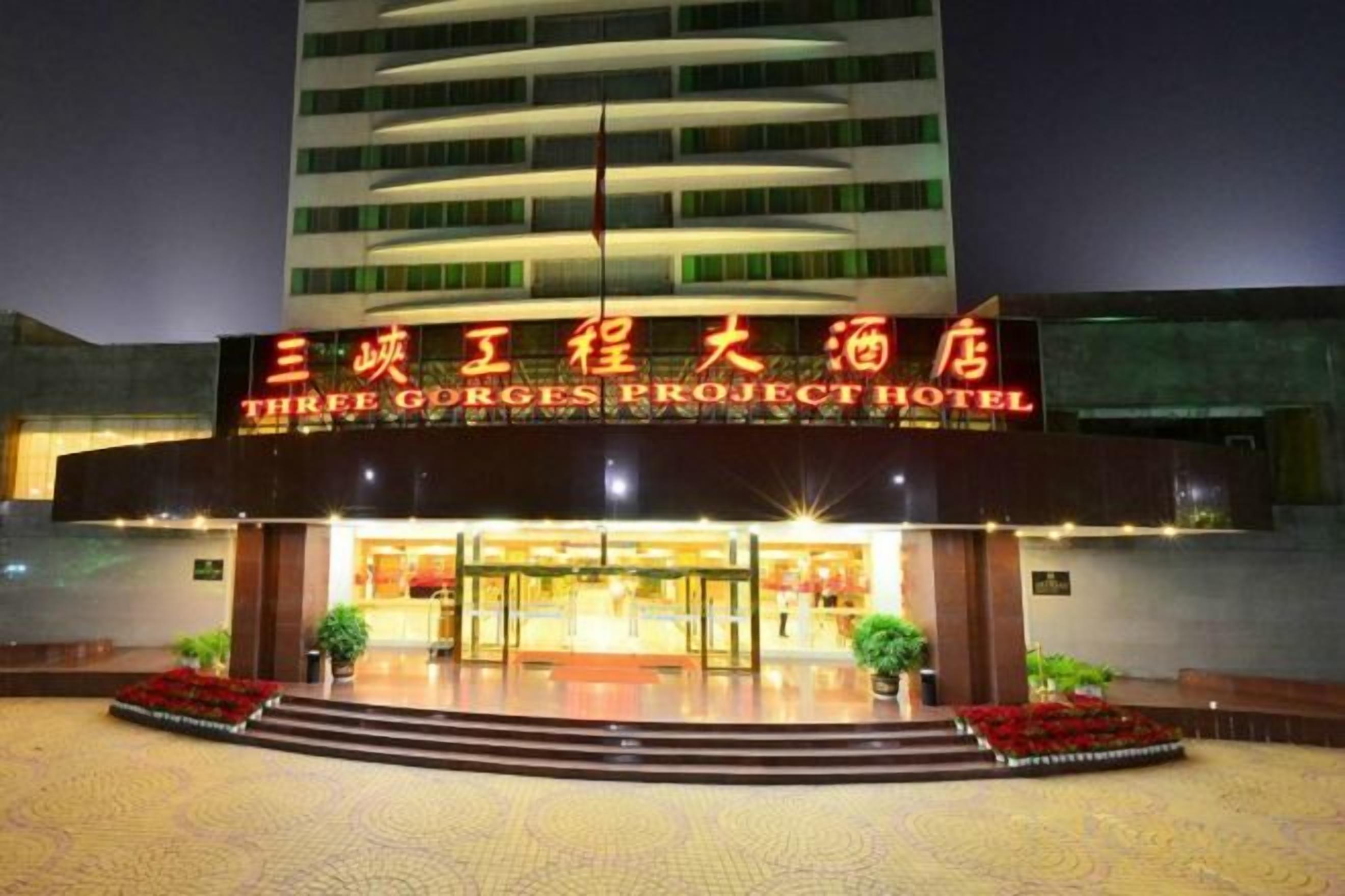 A hotel entrance with a sign in a foreign language, a well-lit facade, and a landscaped area with steps leading up to the entrance.